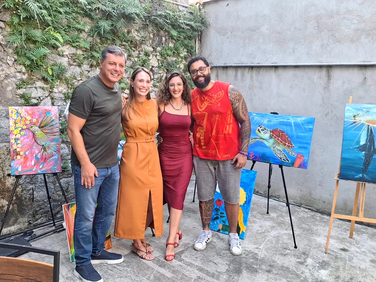 Artista pintora posando para foto entre três fãs com seus quadros expostos ao redor.